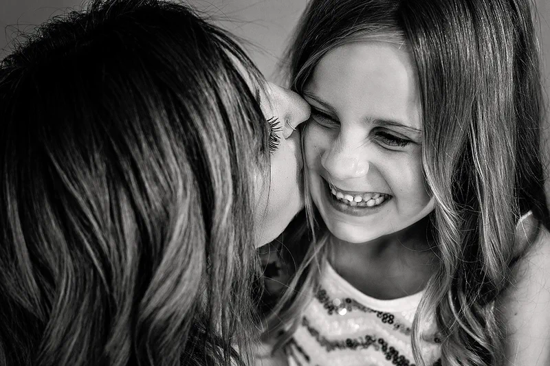 A black and white portrait of a mother kissing her laughing daughter on the cheek