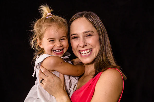 A laughing mother holding her toddler daughter, against a black backdrop